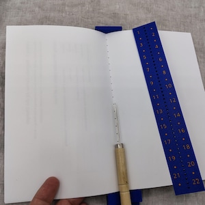 Op de afbeelding: Een open boek met witte pagina's, een blauwe geleider en een priktool. De blauwe geleider heeft de nummers 1-22. De priktool heeft een houten handvat en een metalen naald. Het boek is open op een blanco pagina.