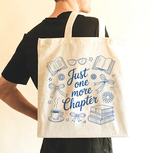 May include: A beige canvas tote bag featuring a blue graphic print with the text Just one more Chapter. The design includes illustrations of open books, a stack of books, eyeglasses, bows, flowers, stars, and a steaming cup of coffee on a saucer.