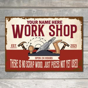May include: A vintage-style metal sign with a red background and white lettering that reads "YOUR NAME HERE WORK SHOP EST. 2021 OPEN 24 HOURS THERE IS NO SCRAP WOOD, JUST PIECES NOT YET USED". The sign features a wood plane, a saw blade, and two hammers.