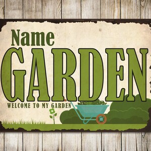 May include: A green and brown metal sign with the text "Name GARDEN" and "WELCOME TO MY GARDEN". The sign features a cartoon image of a wheelbarrow with plants and a small flower.