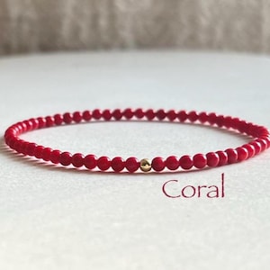 May include: A thin bracelet composed of small, round, vibrant red beads arranged in a continuous circle. A single, polished gold-toned spherical bead is positioned near the center of the strand. The word Coral is printed in red text below the bracelet.