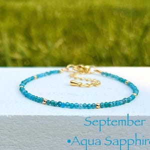 May include: A delicate bracelet featuring small, light blue beads and gold accents. The bracelet is labeled "September Aqua Sapphire" in light blue text. The background is a blurred green.