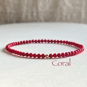 May include: A delicate bracelet composed of small, round, vibrant red beads arranged in a continuous circular strand. A single, polished gold-toned spherical bead is positioned as a central accent. The word Coral is printed in red script below.