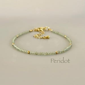 May include: A delicate bracelet featuring small, light green beads and gold-colored accents. The bracelet is strung on a fine chain and has a clasp closure. The word "Peridot" is printed in a serif font.