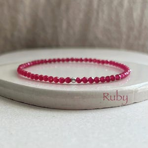 May include: A delicate bracelet composed of small, faceted, vibrant pinkish-red beads arranged in a circular pattern, featuring a single polished silver-toned metal bead centered at the front. The word Ruby is printed in a script font on the surface.