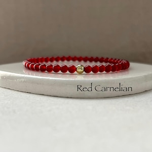 May include: A beaded bracelet featuring small, round, deep red carnelian beads and a single, central, polished gold-tone metal bead. The text "Red Carnelian" is printed on the surface beneath the bracelet, identifying the stone type.