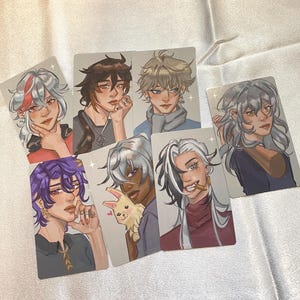 May include: A collection of six illustrated character art cards. Each card features a portrait of a person with unique hairstyles and clothing. The color palette includes shades of purple, red, brown, and gray. The cards are arranged on a white surface.