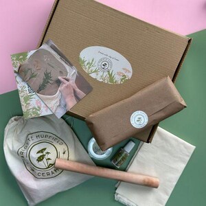 May include: A brown cardboard box containing a white bag, a wooden tool, a small jar, a bottle of liquid, a white cloth, and a brown paper package. The box has a label that says "Create with clay at home" and a logo with the text "Charlotte Hupfield Ceramics".