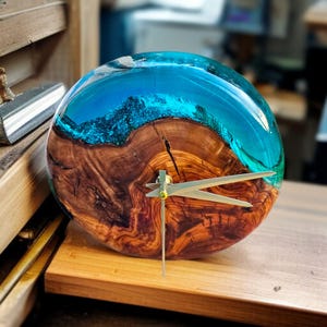 May include: A handcrafted wooden clock with a unique design. The clock face is a combination of wood and blue resin, resembling ocean waves. The clock hands are gold-colored. The clock is sitting on a wooden surface.