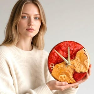 May include: A circular wall clock featuring a combination of natural light-toned wood grain and vibrant red resin. The clock face includes gold-colored hands and a single gold numeral nine positioned on the left side of the wood and resin surface.