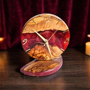 Puede incluir: Un reloj de madera hecho a mano con una esfera redonda y una base curva. La esfera del reloj presenta una combinación de tonos de madera claros y oscuros con un centro de resina rojo intenso. Las manecillas y los números dorados marcan la hora. Es un artículo de decoración.