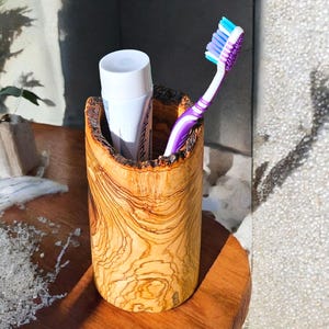 Könnte beinhalten: Ein zylindrischer Zahnbürstenhalter aus hellbraunem Holz mit natürlicher Maserung. Er enthält eine Tube Zahnpasta und eine lila-weiße Zahnbürste. Der Halter steht auf einer Holzoberfläche.