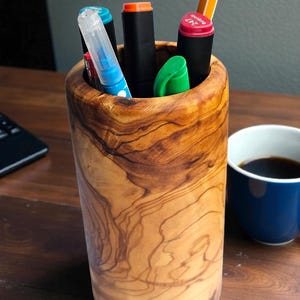 Può includere: Un portapenne cilindrico in legno chiaro, pieno di penne e pennarelli colorati. Il legno mostra un evidente motivo a venature. Una tazza blu di caffè è posizionata vicino su una superficie di legno.