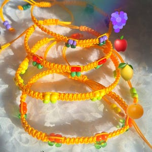 Può includere: Un set di quattro braccialetti in cordino intrecciato giallo con piccole perline di plastica colorate di varie forme. I braccialetti sono realizzati con nodi scorrevoli regolabili e incorporano perline verdi, rosse, viola e gialle.