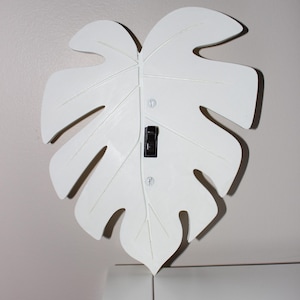 Peut inclure: Interrupteur mural blanc en forme de feuille avec un interrupteur noir. Le couvercle est monté sur un mur de couleur claire. Le design ressemble à une feuille de Monstera, ajoutant une touche de décoration inspirée de la nature.