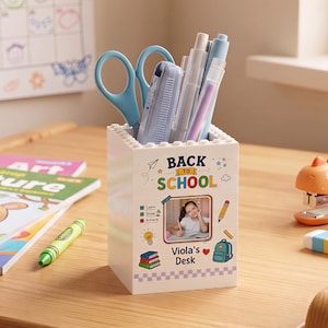 Peut inclure: Un porte-crayons rectangulaire blanc avec une texture de briques de construction sur le bord supérieur. L'avant présente le texte BACK TO SCHOOL, une petite photo d'enfant et le nom Viola's Desk, entourés d'icônes colorées de livres et d'un sac.