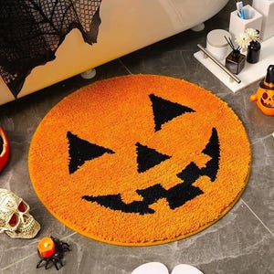 Puede incluir: Alfombra de baño redonda naranja de Halloween con una cara de calabaza. La alfombra tiene ojos triangulares negros y una boca dentada. Un accesorio de baño festivo para la temporada de miedo.