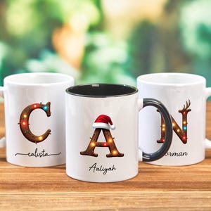 Peut inclure: Trois mugs en c&eacute;ramique blanche avec des motifs de No&euml;l personnalis&eacute;s. Chaque mug pr&eacute;sente une lettre orn&eacute;e de lumi&egrave;res festives et d'un nom. Un mug a un bonnet de P&egrave;re No&euml;l. Les mugs sont sur une surface en bois.