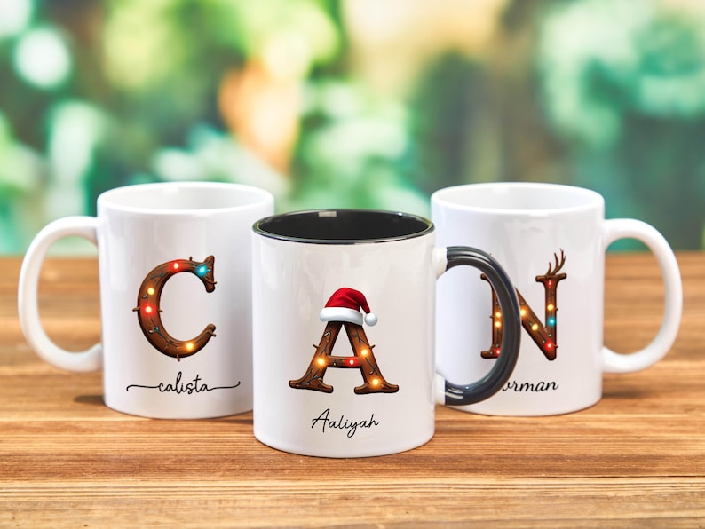 Peut inclure: Trois mugs en c&eacute;ramique blanche avec des motifs de No&euml;l personnalis&eacute;s. Chaque mug pr&eacute;sente une lettre orn&eacute;e de lumi&egrave;res festives et d'un nom. Un mug a un bonnet de P&egrave;re No&euml;l. Les mugs sont sur une surface en bois.