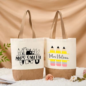 May include: Two canvas tote bags with beige handles and a brown bottom. One bag features the text "MRS. SMITH" with school-themed graphics. The other bag displays "Mrs. Nelson" with pencil graphics. Both bags are on a beige background.