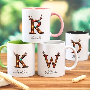 Peut inclure: Quatre mugs en c&eacute;ramique blanche avec des lettres et des noms personnalis&eacute;s. Chaque mug pr&eacute;sente une lettre d&eacute;cor&eacute;e de bois de cerf et de lumi&egrave;res. Les mugs ont des poign&eacute;es et des int&eacute;rieurs de couleurs diff&eacute;rentes, dont rose, vert et noir.