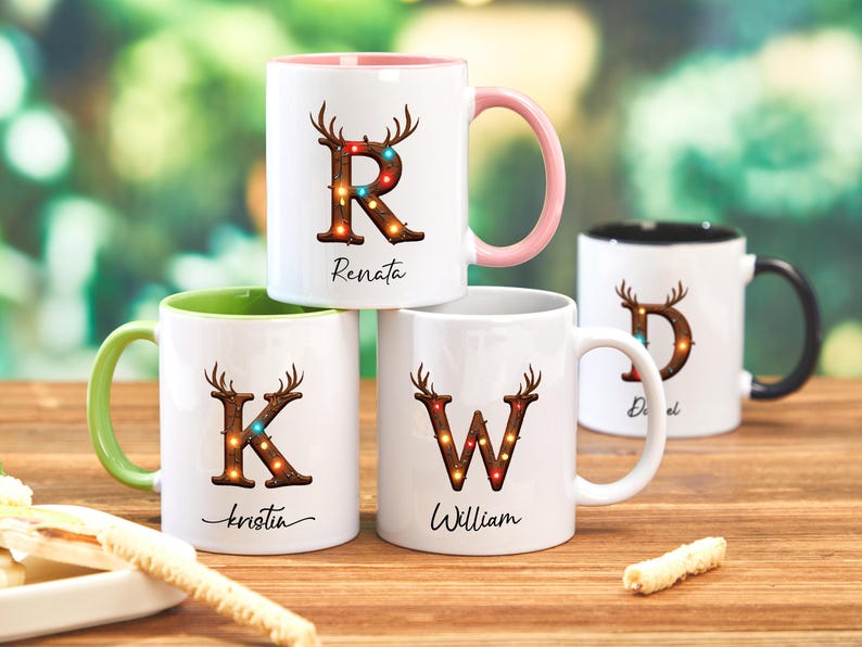 Peut inclure: Quatre mugs en c&eacute;ramique blanche avec des lettres et des noms personnalis&eacute;s. Chaque mug pr&eacute;sente une lettre d&eacute;cor&eacute;e de bois de cerf et de lumi&egrave;res. Les mugs ont des poign&eacute;es et des int&eacute;rieurs de couleurs diff&eacute;rentes, dont rose, vert et noir.