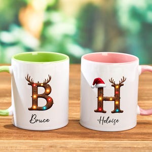 Peut inclure: Deux mugs en c&eacute;ramique blancs avec des poign&eacute;es et int&eacute;rieurs color&eacute;s. L'un a une poign&eacute;e et un int&eacute;rieur verts, l'autre une poign&eacute;e et un int&eacute;rieur roses. Les deux mugs pr&eacute;sentent une lettre d&eacute;cor&eacute;e de bois de cerf et de lumi&egrave;res, avec un nom en dessous.