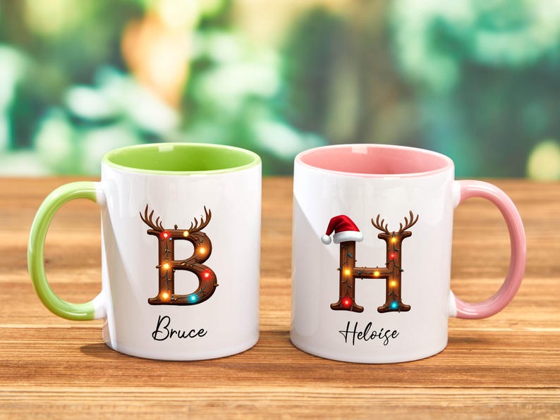 Peut inclure: Deux mugs en c&eacute;ramique blancs avec des poign&eacute;es et int&eacute;rieurs color&eacute;s. L'un a une poign&eacute;e et un int&eacute;rieur verts, l'autre une poign&eacute;e et un int&eacute;rieur roses. Les deux mugs pr&eacute;sentent une lettre d&eacute;cor&eacute;e de bois de cerf et de lumi&egrave;res, avec un nom en dessous.