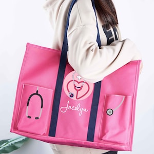 May include: A pink tote bag with navy blue straps and pockets. The bag features a stethoscope and heart design with the name "Jocelyn". The bag is designed for medical professionals.