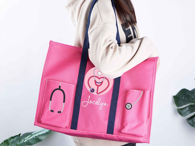 May include: A pink tote bag with navy blue straps and pockets. The bag features a stethoscope and heart design with the name "Jocelyn". The bag is designed for medical professionals.