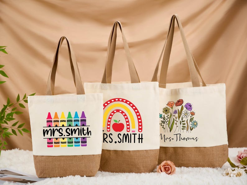 May include: Three canvas tote bags with brown bottoms and handles. The bags feature colorful designs: crayons with "Mrs. Smith", a rainbow with an apple and "RS. Smith", and a floral design with "Mrs. Thomas".