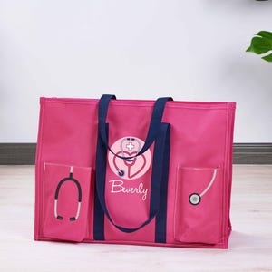 May include: A pink tote bag with navy blue handles and accents. The bag features a stethoscope design with the name "Beverly" in script. Two pockets on the front display stethoscope graphics. A practical and stylish bag for medical professionals.