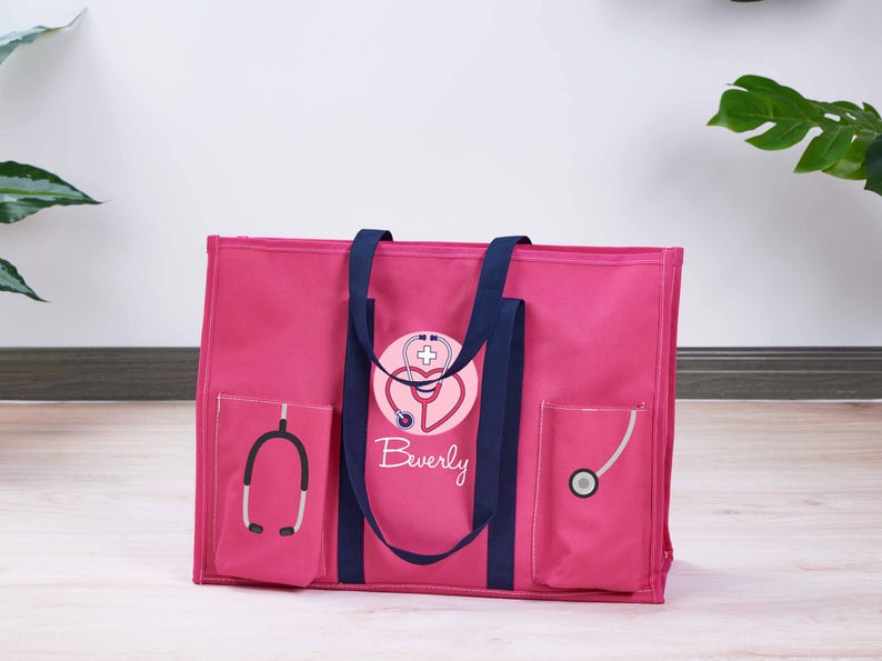 May include: A pink tote bag with navy blue handles and accents. The bag features a stethoscope design with the name "Beverly" in script. Two pockets on the front display stethoscope graphics. A practical and stylish bag for medical professionals.