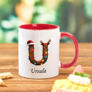 Peut inclure: Mug en c&eacute;ramique blanche avec int&eacute;rieur et anse rouges. La lettre "U" est d&eacute;cor&eacute;e d'un bois de renne et d'un bonnet de P&egrave;re No&euml;l, et orn&eacute;e de lumi&egrave;res color&eacute;es. Le nom "Ursula" est imprim&eacute; en dessous.