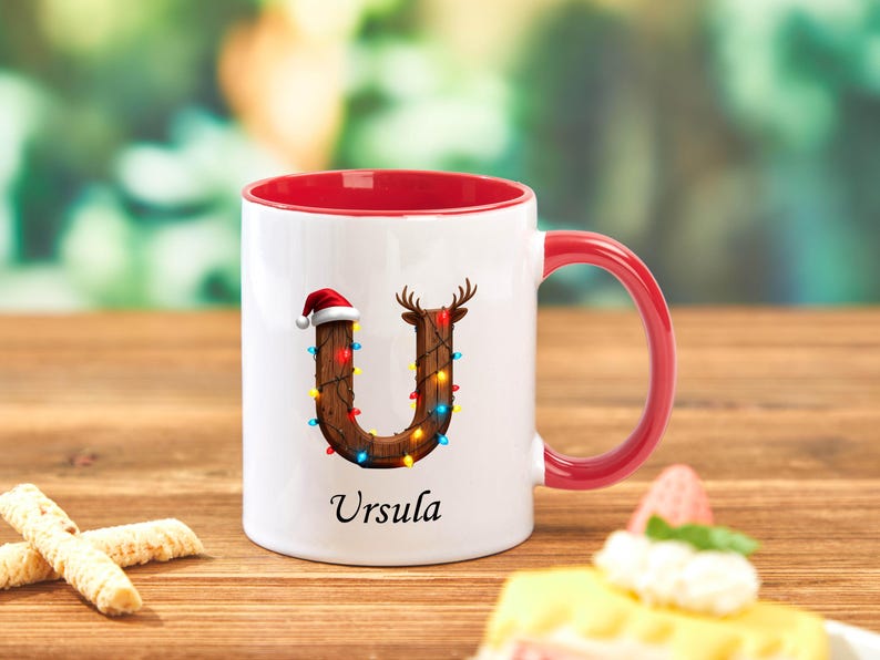 Peut inclure: Mug en c&eacute;ramique blanche avec int&eacute;rieur et anse rouges. La lettre "U" est d&eacute;cor&eacute;e d'un bois de renne et d'un bonnet de P&egrave;re No&euml;l, et orn&eacute;e de lumi&egrave;res color&eacute;es. Le nom "Ursula" est imprim&eacute; en dessous.