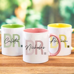 Peut inclure: Trois tasses en céramique blanche avec des bords et des poignées de couleur. Les tasses ont les lettres "DR." imprimées sur elles avec un nom sous chaque ensemble de lettres. Les tasses sont sur une surface en bois. Les noms sont Calista, Nicholas et Antonia.