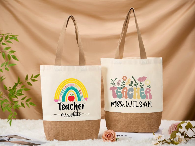 May include: Two canvas tote bags with beige handles and a brown bottom. One bag features a rainbow design with an apple and the text "Teacher mrs. White." The other bag has the word "Teacher" with floral accents and the text "mrs. Wilson."