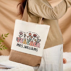 May include: A canvas tote bag with a beige top and a brown bottom. The bag features the word "TEACHER" in pastel colors, with floral accents and the name "MRS. WILLIAMS" in black. The bag has beige handles.