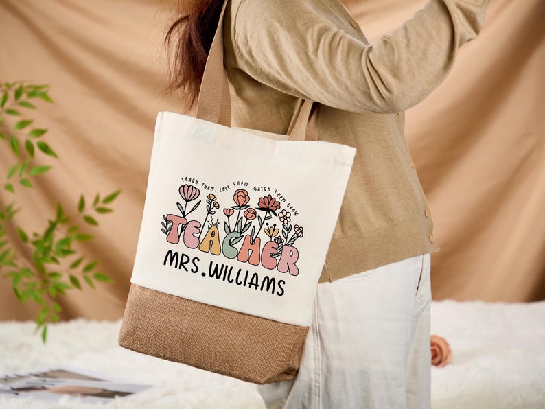 May include: A canvas tote bag with a beige top and a brown bottom. The bag features the word "TEACHER" in pastel colors, with floral accents and the name "MRS. WILLIAMS" in black. The bag has beige handles.