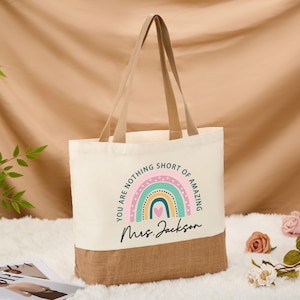 May include: Cream-colored tote bag with a rainbow design and the text "YOU ARE NOTHING SHORT OF AMAZING" and "Mrs. Jackson". The bottom of the bag is a natural burlap color, with matching handles.