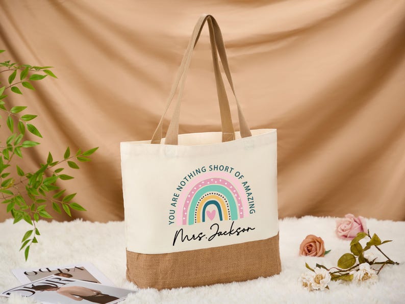 May include: Cream-colored tote bag with a rainbow design and the text "YOU ARE NOTHING SHORT OF AMAZING" and "Mrs. Jackson". The bottom of the bag is a natural burlap color, with matching handles.