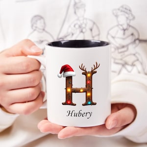 Peut inclure: Mug en c&eacute;ramique blanche avec int&eacute;rieur noir, orn&eacute; d'un motif festif. Le motif comprend la lettre "H" en bois orn&eacute;e de guirlandes lumineuses, de bois de cerf et d'un bonnet de P&egrave;re No&euml;l. Le nom "Hubery" est imprim&eacute; en dessous.