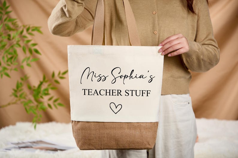 May include: Cream-colored tote bag with the text "Miss Sophia's TEACHER STUFF" in black script, a heart symbol, and a brown bottom. The bag has beige handles.