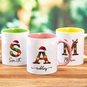 Peut inclure: Trois mugs en c&eacute;ramique blanche avec des int&eacute;rieurs et des poign&eacute;es color&eacute;s. Chaque mug pr&eacute;sente une lettre d&eacute;cor&eacute;e de guirlandes lumineuses et d'un bonnet de P&egrave;re No&euml;l. Les mugs portent les noms "Smith", "ashley" et "Maria".