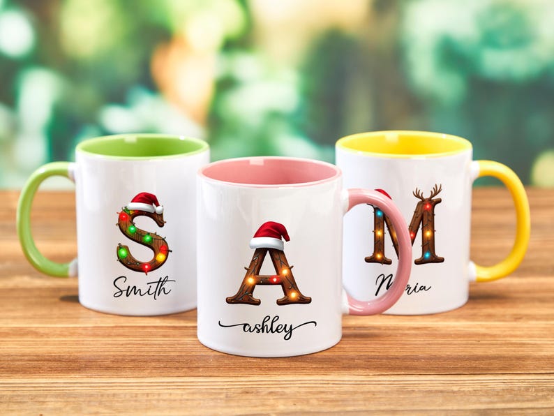 Peut inclure: Trois mugs en c&eacute;ramique blanche avec des int&eacute;rieurs et des poign&eacute;es color&eacute;s. Chaque mug pr&eacute;sente une lettre d&eacute;cor&eacute;e de guirlandes lumineuses et d'un bonnet de P&egrave;re No&euml;l. Les mugs portent les noms "Smith", "ashley" et "Maria".