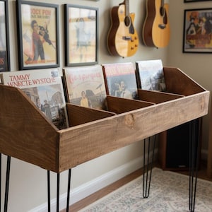 May include: A wooden record storage unit with black hairpin legs. The unit is filled with vinyl records. The wood has a dark stain. Framed posters and guitars hang on the wall in the background.