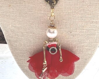 May include: A whimsical necklace featuring a doll pendant. The doll has a red tulle skirt, gold-toned limbs, and a pearl head. The necklace chain includes red and white beads and gold-toned metal accents. The pendant also has a small purse.