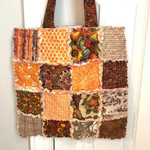 May include: A tote bag constructed from a patchwork of quilted fabric squares featuring various autumn-themed patterns in shades of orange, brown, yellow, and cream. The bag has frayed seams between each square and two long, dark brown patterned fabric handles.