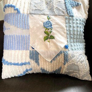 May include: A square throw pillow featuring a patchwork design of white and light blue textured fabrics. The center panel is a smooth white fabric square with an embroidered blue rose with green leaves and a stem, set in a diamond orientation.