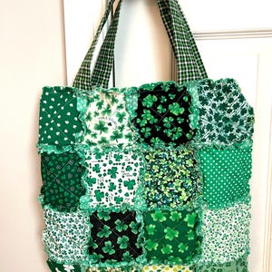 May include: A patchwork tote bag constructed from various green and white fabric squares featuring shamrock patterns, polka dots, and floral motifs. The bag has frayed seams between the squares and two green and white gingham handles.